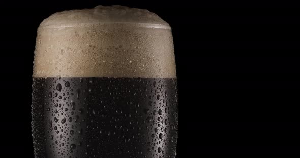 Glass of black beer on a black background. Beer sways in the glass, bubbles and foam rise.