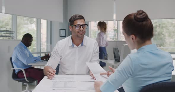 Back View of Female Manager Sitting at Desk and Consulting Indian Male Client in Office