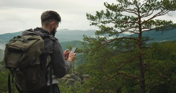 A Man is Standing on Top of a Mountain and Texting a Message on His Smartphone