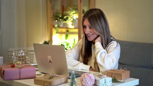 Happy and Smiling Young Woman Wrapping and Preparing Christmas Gifts Using Laptop and Having Online