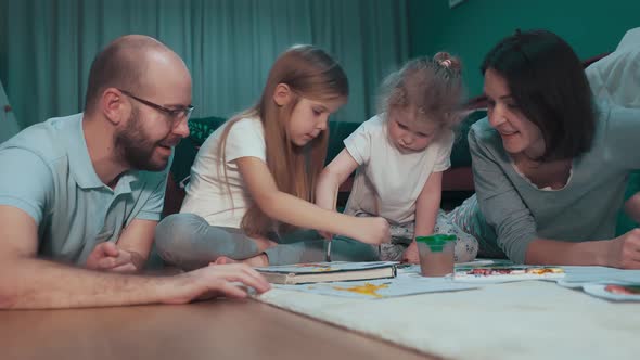 Mother and father teach their two daughters draw pictures in the living room on the floor alt