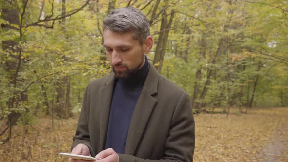 Handsome Caucasian Man Standing in the Autumn Forest with Tablet and Looking Around