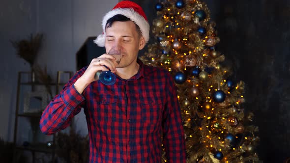 Young Man in Santa Hat Drinks Champagne and Grimaces From Alcohol on Background of Christmas Tree in