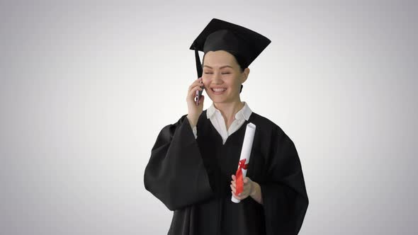 Emotional Female Student in Graduation Robe Talking on the Phone Holding Diploma on Gradient