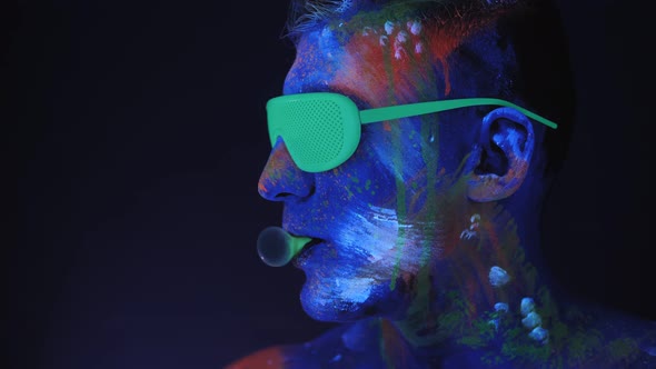 A Man with UV Drawings and Green Neon Glasses Inflates a Bubble of Bubblegum