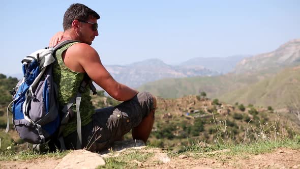 Man with backpack sits on edge of a cliff in mountains and enjoys beautiful panorama view