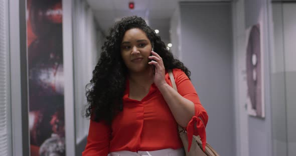 Smiling biracial businesswoman talking on smartphone walking down corridor in modern office