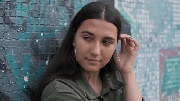 Thoughtful Young Brunette Girl Runs Her Hand Through Her Hair and Leans on the Wall with Graffiti