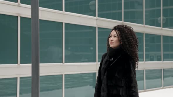 A Curlyhaired Stylish Middleaged Brunette Woman in a Black Fur Coat Walks Alone in the City on a