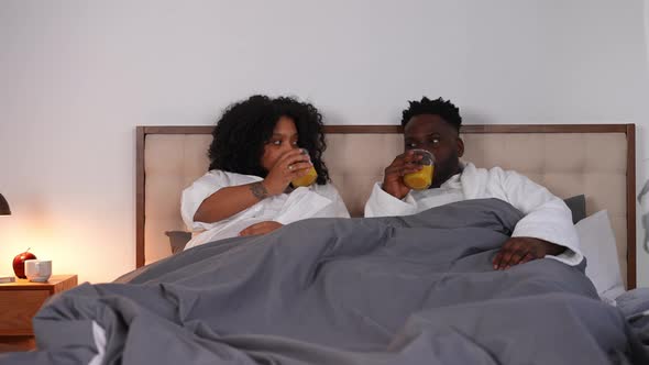 Carefree Happy African American Young Couple Talking Drinking Orange Juice Lying in Bed in the