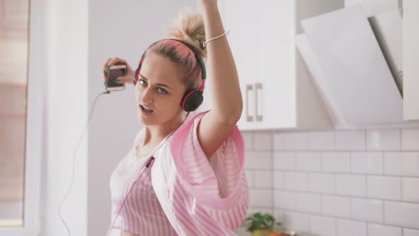 Happy Young Woman with Pink Hair Dancing in Kitchen Wearing Pink Pajamas and Listening to Music with