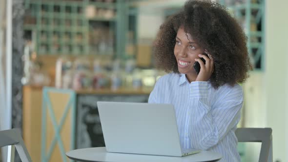 African Woman with Laptop Talking on Smartphone in Cafe 