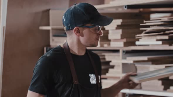 Man in a Carpentry Workshop Takes a Wooden Piece From the Shelf and Blowing Dust From It Looking in