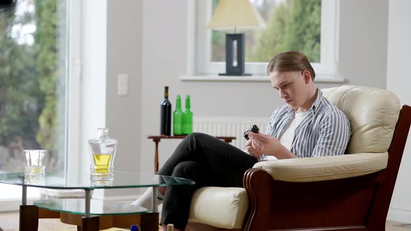 Wide Shot of Depressed Young Man with Gun Sitting on Armchair with Alcohol on Table