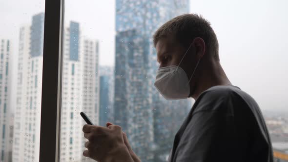 Side View of a Man in a Face Mask Using His Smartphone Sitting Alone By the Window