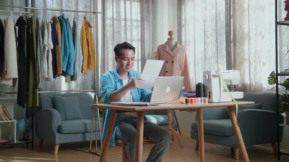 Asian Male Designer With Sewing Machine Designing Clothes On Paper While Working With Laptop