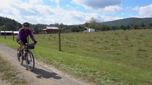 Mature woman with sunglasses biking on a gravel road with a farm in the distance.