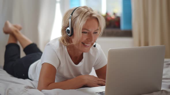 Cheerful Grandmother in Headset Talking with Relatives Via Laptop Lying on Bed