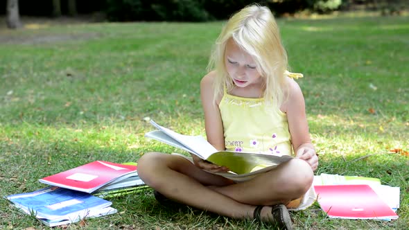 Little Cute Smart Girl Sits in the Park Surrounded By Many Workbooks - She Read One and Study