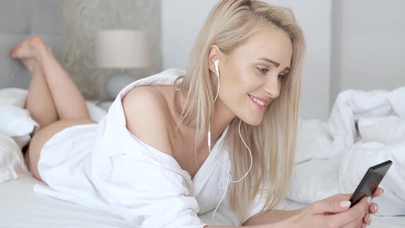 Beautiful, Smiling Blond Woman Lying in White Bed and Using a Smartphone