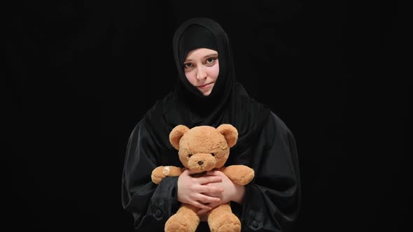 Middle Shot Portrait of Shy Muslim Woman in Hijab Posing at Black Background with Teddy Bear