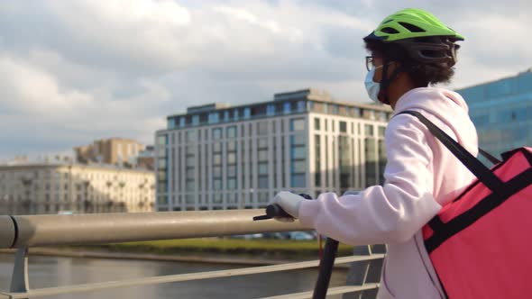 African Delivery Man on Scooter Standing on Bridge Putting Off Safety Mask and Enjoying Fresh Air