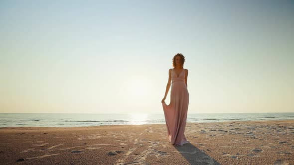 Cute Woman in Long Dress Walking on Sand at you Against Sunset Sea Background