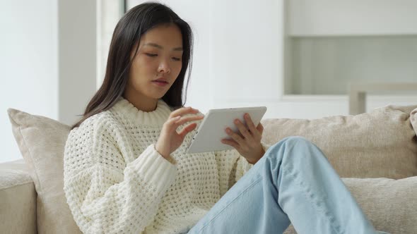 Asian Woman Uses a Tablet While Sitting on a Home Sofa in Her Living Room