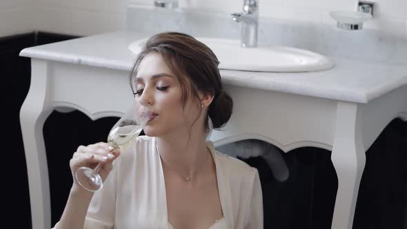 Bride in White Boudoir Dress Sitting on Floor in Bathroom at Home Apartment with Glass of Champagne
