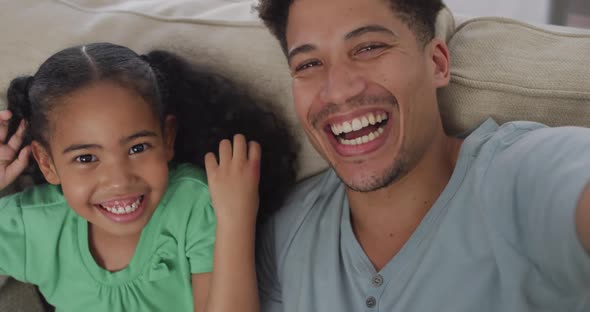 Happy biracial father and daughter sitting on sofa taking selfies