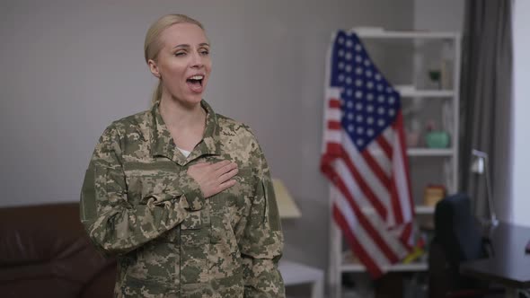 Proud Young Woman in Camouflage Uniform Singing National Anthem Standing Indoors with USA Flag at