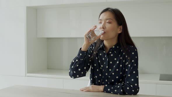 Young Asian Woman Drinks Water From a Glass Sitting in the Kitchen