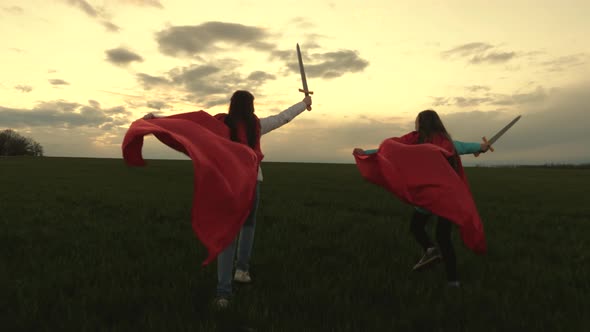 Girls in Red Cloaks Run with Swords in Hand Across Field Playing Medieval Knights. Children Fight