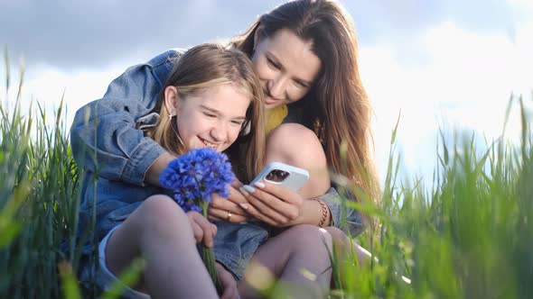 Young Mother and Daughter Relaxing in Nature in the Park They Have Fun Laughing Using a Smartphone