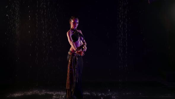 A Woman Dances an Oriental Dance on a Black Background in the Pouring Rain in the Studio