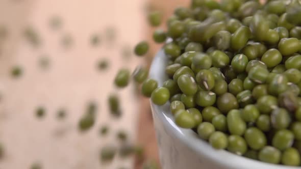 Raw green mung beans fill a white cup and scatter across a wooden surface in slow motion