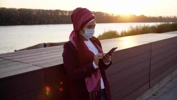 Young Muslim Woman in Medical Mask with Phone Outdoors at Sunset