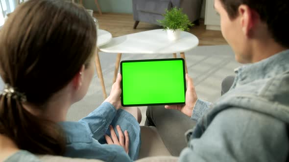 Over The Shoulder View Young Couple at Home Sitting on a Sofa Using With Green Mock-up Screen Tablet