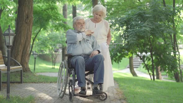 Wide Shot of Sad Beautiful Caucasian Senior Woman Supporting Disabled Man Sitting in Wheelchair. Old