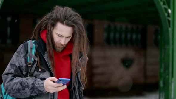 Trendy Man with Dreadlocks is Using Smartphone on Street in Winter Swiping and Scrolling Display