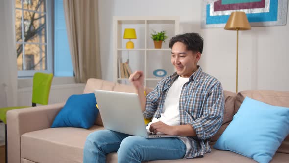 Young Asian Man with Raised Arms Celebrating Victory Sitting on Couch and Using Laptop at Home