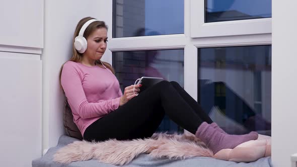 Young Woman Sits on Windowsill Near Large Window Wearing Big White Headphones Watching Movie on