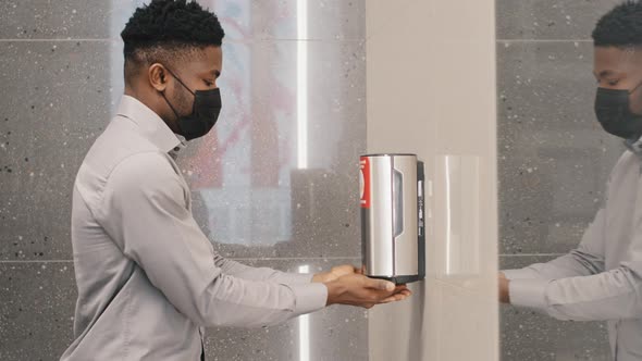 Young African American Man in Protective Mask Stands in Public Toilet Dries Hands Under Jet of Hot