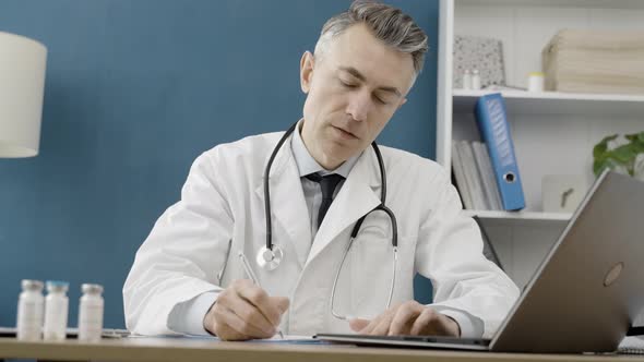 Senior male doctor with a stethoscope in a lab coat sitting at the desk