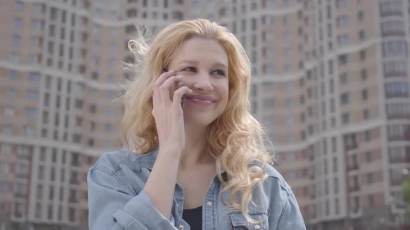 Close-up Face of Pretty Smiling Confident Blond Woman Talking By Cellphone in Front of Skyscraper