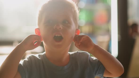 Little Boy with Open Mouth Closes Ears By Fingers in Room