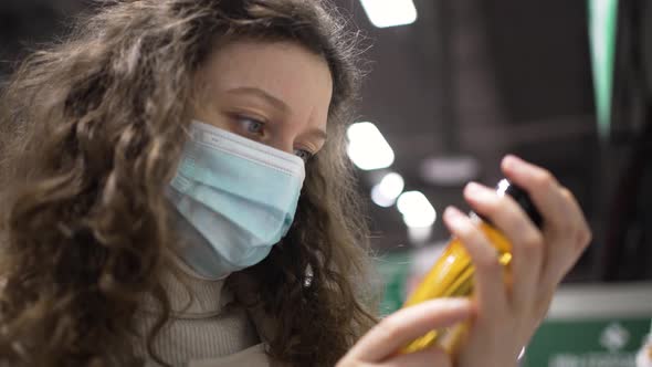 Woman Customer in a Medical Mask Reads the Composition on a Bottle of Oil in a Supermarket Closeup