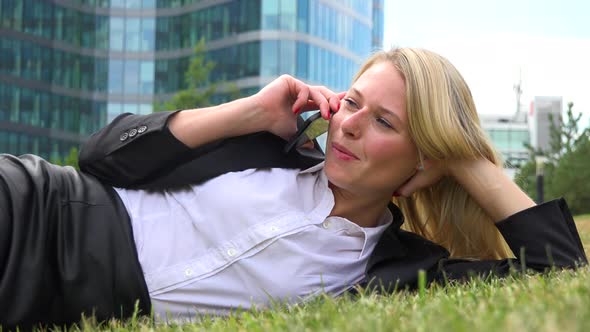 A Young Beautiful Businesswoman Lies in Grass in a Park and Talks on a Smartphone - Closeup