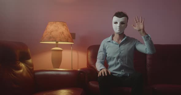 Anonymous man with mask greets with his hand while sitting on the red sofa at home