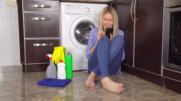 a woman is sitting on kitchen floor with her phone. Bottles with cleaning agent